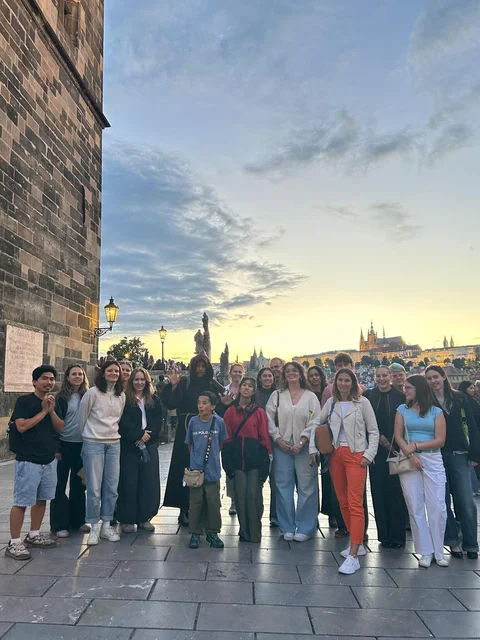 Ghost Tour guests with a hooded guide beside the Charles Bridge tower at sunset, with Prague Castle in the background.