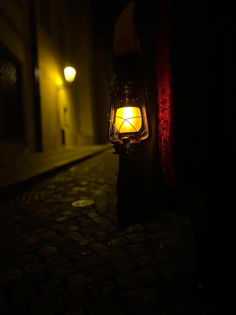A glowing lantern held beside a dark cloak on a cobbled Prague street at night.