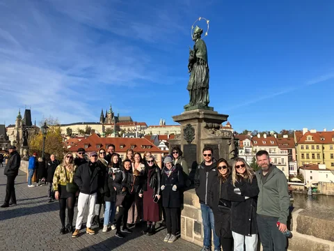 Travellers beside a statue on Charles Bridge, with the Vltava and red-roofed buildings behind them.