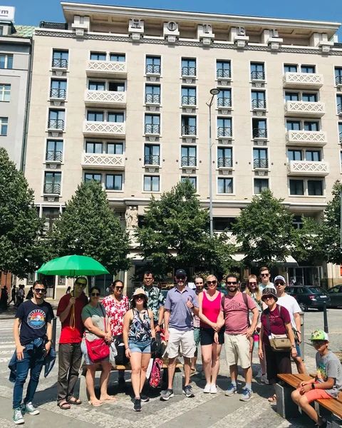New Town tour guests with a green umbrella beside trees and benches, with city buildings behind them.